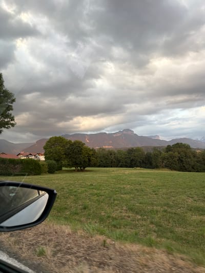 The image shows a rural landscape with a cloudy sky. In the foreground, a car's side mirror is visible on the left, reflecting a portion of the sky and a tree. Beyond the mirror, a grassy field stretches into the midground. A line of trees borders the field, and behind them, houses with red roofs and white walls are visible. In the background, a mountain range is silhouetted against the sky, with some areas illuminated by sunlight breaking through the clouds. The scene appears to be a view from a moving vehicle. The location is identified as Groisy, France.
