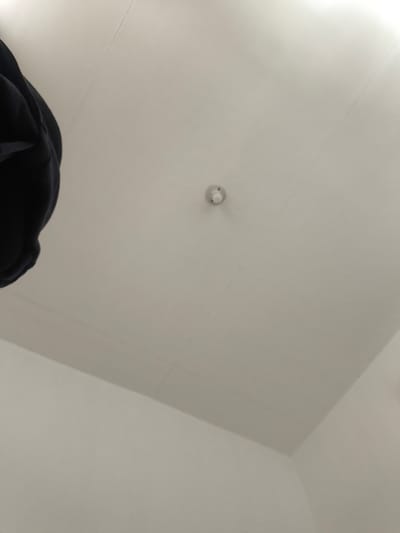 The image shows a ceiling with a smoke detector. A dark garment is visible in the upper left corner of the frame, partially obscuring the ceiling. The ceiling appears to be white and made of panels, with faint lines indicating seams. The smoke detector is small and round, with a white central button or indicator light. The scene is indoors, with the white walls suggesting a room. The lighting is neutral, and no specific time of day or weather conditions can be determined. There are no discernible text elements in the image. The overall impression is that of a common indoor setting, likely a home or office, with functional elements like a smoke detector.