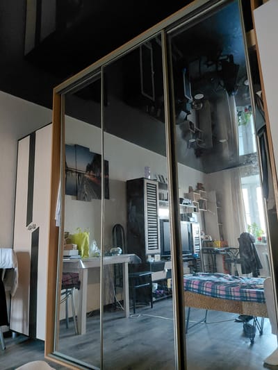 This media file captures a reflection of a room, likely a bedroom or living area, in a large, multi-paneled mirrored surface, possibly a wardrobe or sliding door. The image is oriented vertically, with the reflection appearing sideways.
The reflected room features a bed positioned to the right, covered with a blue, pink, and white plaid blanket. To the left of the bed, a collection of furniture includes shelves displaying various items, a desk or table with what appears to be a laptop or monitor, and chairs. Clothes are visible draped over some furniture or a rack. A light-colored plastic bag hangs near the desk area. On a wall, three framed pictures are visible, one depicting a city bridge at night. In the background of the reflection, a window with a white frame allows natural light into the reflected room, and some greenery is visible outside, suggesting an outdoor view or another room with plants. The floor of the reflected room is dark wood or laminate, and the walls are light-colored.
The mirror itself has a light wooden frame along the top and bottom edges, with metallic horizontal strips dividing the glass panels. The foreground, representing the room from which the photograph was taken, is mostly out of focus and shows dark walls, possibly black or dark gray, and a piece of white furniture with black horizontal stripes.
No people are visible in the image. There is no specific activity depicted, but the arrangement of furniture suggests a functional living space. The lighting from the reflected window indicates it is likely daytime. No legible text is visible. The location context is Nadym, Russia, but no specific visual cues within the image confirm this.