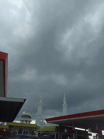 The image depicts a mosque with two tall white minarets and a domed structure, likely located in the city of Meunasahaluedrienlhoksukon Barat, Indonesia. A portion of a building with a red and grey facade is visible on the left. In the foreground, a covered structure with a red roof, possibly a gas station canopy, is present. The sky is overcast with dark, heavy clouds, suggesting impending rain or a storm. Several vehicles are partially visible in the lower part of the frame, and some structures with yellow and white elements are situated near the mosque. The scene appears to be a street-level view of an urban or semi-urban environment.