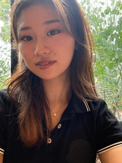 The image shows a close-up portrait of a young woman with long brown hair. She is wearing a black polo shirt with white stripes on the collar and sleeves. The shirt has a small gold cross necklace and an embroidered logo on the left breast. The woman is looking directly at the camera, with a neutral expression on her face. Her face is illuminated by natural light coming from behind the camera, casting a subtle shadow on the right side of her face. The background is a blurry out-of-focus shot of green foliage, possibly a garden, with a window visible on the left side of the image. The location is Takarazuka, Japan, a city known for its beautiful gardens and natural scenery. The coordinates provided correspond to a residential area in Takarazuka, near the Takarazuka Grand Theater, a famous musical theater. The image appears to have been taken indoors, possibly near a window, as the natural light is soft and diffused, creating a sense of intimacy. The woman's attire suggests she may be enjoying leisure time in a relaxed setting.
