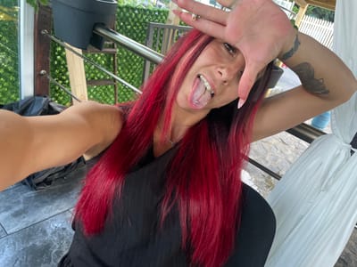 This media file shows a close-up, upside-down, selfie-style shot of a woman with striking red hair and a black top. She is outdoors, with a blurry green background suggesting foliage and possibly a balcony or outdoor seating area with metal railings. A white draped fabric, possibly a curtain or canopy, is visible on the left. The woman is sticking out her tongue, which has a silver piercing. Her right hand is partially covering her face. She has long, light-colored fingernails and a tattoo on her left arm. The lighting suggests it is daytime, possibly bright, as indicated by the shadows and the overall illumination. The overall mood appears playful or provocative, given her expression and the pose.