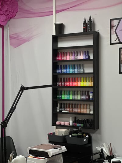 This image, taken in Kuda, Russia, depicts a professional nail salon workspace, meticulously organized and equipped. The central focus is a black, wall-mounted shelving unit filled with numerous small, rectangular bottles, likely nail polishes or gel colors. These bottles are neatly arranged in distinct color gradients, showcasing purples, blues, greens, yellows, reds, and various shades of brown. Many of the bottles prominently feature the text "BH." On the upper left side of the shelving unit, two black squeeze bottles are positioned.
To the right of the shelving unit, a collection of nail salon tools and accessories is visible. This includes a black organizer holding additional bottles and a white and rose gold/bronze nail dust collector/fan, which is labeled "LUM-AUS." Below the shelving unit, a black articulated arm, resembling a task lamp or a stand, extends into the foreground.
The background features a light-colored wall, possibly white or light grey, adorned with a soft, swirling pink pattern on the left side. A large, decorative pink artificial flower is situated in the lower-left corner of the frame. Above the shelving unit, two framed items are mounted on the wall; one is a certificate with the partial text "CERT" visible, and the other is an indistinct framed picture. No individuals are present in the image, but the arrangement of specialized equipment and products clearly indicates an area dedicated to nail services.