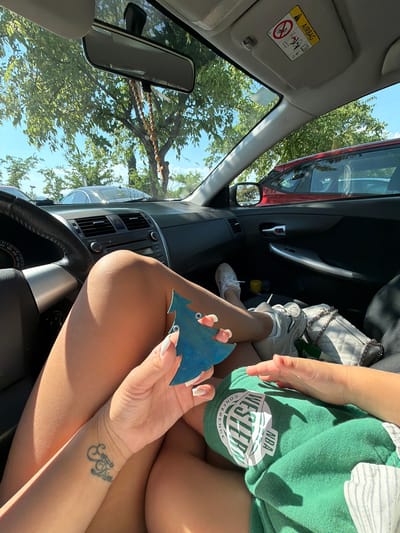 The image shows the interior of a car parked outdoors. In the foreground, a person's legs are visible, with one hand holding a blue tree-shaped ornament. The person is wearing white sneakers and a green t-shirt. A tattoo is visible on the person's wrist. In the background, trees and other cars can be seen through the car windows. The sun is shining, indicating it is daytime.