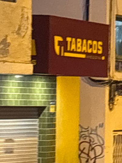 The image captures a close-up view of a shop front in Calp, Spain. The primary focus is a burgundy sign with yellow lettering that reads "TABACOS," indicating a tobacco shop. To the left of the sign, a textured, light-colored wall is visible, possibly stone or stucco, with a faint leafy pattern. Below the sign, a green tiled facade with horizontal white stripes runs across the width of the image. A metallic roll-up door with horizontal slats is partially visible beneath the tiled section. To the right of the "TABACOS" sign, a bright yellow pillar stands, and further to the right, a glimpse of a building with a window and vertical blinds can be seen. The bottom right portion of the image shows a light-colored pillar or wall with black graffiti. The overall scene suggests an urban commercial area, likely at dusk or night, given the lighting conditions.