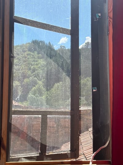 A close-up view through a window screen shows a sunlit outdoor scene. The screen is attached to a wooden window frame, and a section of a red curtain is visible on the right side of the frame. Outside the window, a lush, green hillside is covered with trees. The sky is blue with a few white clouds. In the foreground, the rooftops of buildings are visible, with terracotta tiles. A thin white cable extends from the bottom right corner of the frame, disappearing behind the screen. Two small, square colorful stickers are adhered to the dark vertical frame on the right. The image appears to be taken from inside a building in Shipkovo, Bulgaria, looking out.