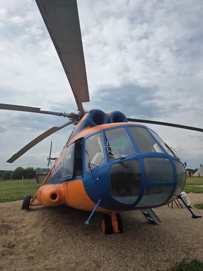 This is a low-angle, full-frame photograph of a blue and orange helicopter parked on a gravel surface. The helicopter is positioned in the center of the frame and occupies the majority of the image. The front of the helicopter, with its distinctive blue cockpit and large windows, faces the viewer. Two large rotor blades extend upwards and outwards from the top of the helicopter. The helicopter's body is predominantly orange, with blue accents around the cockpit and engine cowlings. One of the cabin doors is open. The helicopter is resting on three landing gear wheels, one of which is partially obscured by the gravel. In the background, there is a green grassy field with trees and some small wooden structures, including what appears to be a small log cabin and a conical tent-like structure. The sky is overcast with dramatic grey and white clouds. The ground beneath the helicopter is composed of loose gravel and dirt. The overall scene suggests an outdoor display or static exhibition of the aircraft, likely in a rural setting. The location in Mashino, Russia is not visually confirmed by the image itself, but assumed from the prompt.