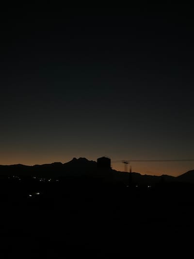 The image depicts a twilight or pre-dawn panoramic view over San Buenaventura, Mexico. The sky transitions from dark grey-black at the zenith to a muted orange-amber glow along the horizon. In the midground, a mountain range is silhouetted against the horizon, with a prominent, jagged peak situated towards the center-left. To the right of this peak, a large, dark, cylindrical structure, possibly a water tower or industrial tank, stands silhouetted. Further to the right, a utility pole with horizontal overhead lines is partially visible. Scattered small, bright points of light are present in the lower left midground, indicative of distant city or street lighting. The foreground is entirely obscured in darkness, rendering any immediate details indistinguishable. No people, specific actions, or interactions are visible.