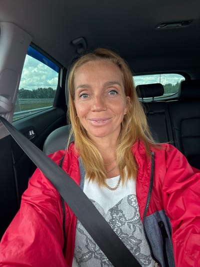 A woman with blonde hair and freckles is taking a selfie in a car. She is wearing a red jacket over a white t-shirt with a lace-like pattern. A seatbelt is across her chest. The interior of the car, including black seats and a window with a view of trees and clouds, is visible. The image was taken in Ozernoye-2, Russia.