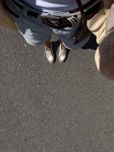 The image, taken from a high angle, shows a person standing on a paved surface that appears to be asphalt. The person is wearing light-wash denim shorts, a white t-shirt, a brown belt with a silver buckle, and light-colored sneakers with yellow laces. A beige jacket is visible on the person's left side, and a brown bag is hanging from their right shoulder. A single white cigarette butt rests on the pavement near the person's feet. The scene is outdoors, under bright sunlight, casting shadows. The background consists of the dark asphalt surface, which fills most of the frame, suggesting an urban or public outdoor location. Without specific landmarks or context, it is impossible to definitively place this scene in Kolašin, Montenegro.