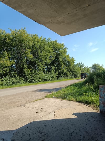 The image shows an outdoor scene on a sunny day, likely in the city of Posad, Russia. In the foreground, there is a concrete structure casting a shadow on a lighter concrete surface. A paved road runs diagonally from the left towards the right of the image, bordered by lush green trees and bushes on the left and more grass and foliage on the right. A dark-colored van is visible on the road, moving away from the viewer and towards the right side of the frame. The sky is clear and blue with a few wispy white clouds. A portion of a weathered, painted wall is visible on the far right edge of the image.