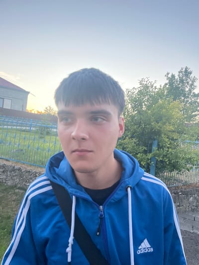 A young male, appearing to be in his late teens or early twenties, stands outdoors, facing slightly to his left with his gaze directed away from the camera. He has short, dark, somewhat messy hair and light-colored eyes. He is wearing a bright blue Adidas hooded jacket with white stripes on the sleeves and a black strap across his chest.

The background suggests a suburban or rural setting. To the left, a portion of a building with a reddish-brown roof is visible, along with a light blue metal fence. Behind the fence, there is a grassy area. To the right of the individual, there are lush green trees with dense foliage. The sky above is a pale blue, indicating it might be late afternoon or early morning, possibly during sunset or sunrise, given the soft lighting. The ground in the foreground appears to be a mixture of gravel and stone. The image's composition and lighting imply it was taken with a mobile phone camera. There is no specific indication of the location being the city of Tovstoluh, Ukraine, within the image itself.