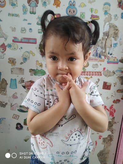 A young child with pigtails stands in front of a busy wall. The girl is wearing a white shirt with a dinosaur pattern, and her fingers are in her mouth. The wall behind her features various cartoon stickers and peeling paint, creating a cluttered and slightly distressed backdrop. The text "SHOT ON REDMI 7 AI DUAL CAMERA" is visible in the bottom left corner.