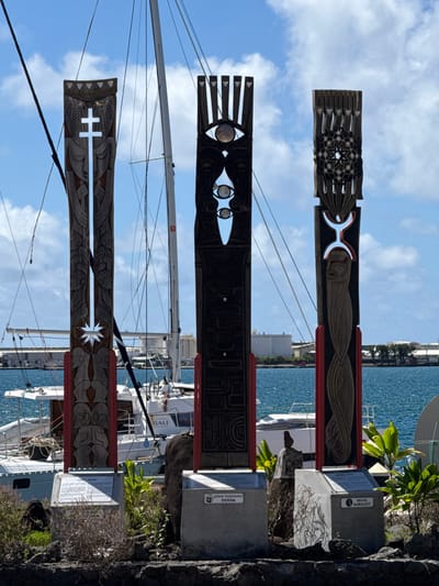 This image depicts three tall, intricately carved wooden sculptures positioned along the waterfront in Papeete, France, specifically within its port or marina area. The sculptures stand upright on individual concrete bases atop rocky ground, interspersed with green foliage.

The leftmost sculpture is a dark brown, vertically oriented plank featuring detailed carvings. Its apex displays triangular patterns framing a stylized face. Below this, a prominent, elongated void shaped like a Marquesan cross dominates the upper-middle section. The lower portion reveals intertwined, anthropomorphic figures. Red accents are present along its lower sides, and it rests on a concrete base with an informational plaque.

The central sculpture, also a dark brown wooden plank, exhibits a series of carved elements. Its uppermost section features five upward-pointing prongs or fingers. Below this, a large stylized eye with a reflective circular orb is carved, followed by a vertical, tear-drop shaped void containing two smaller, similar eyes with orbs. This sculpture also has red accents along its lower sides and stands on a concrete base with an informational plaque identifying "JOHN TARDANEI DOOM."

The rightmost sculpture is another dark brown, carved wooden plank. Its upper portion showcases a dense, circular geometric pattern resembling a star or rosette. Directly beneath this, a prominent void forms the shape of a stylized human figure with outstretched arms, situated above an inverted U-shape. The lower part features swirling or wave-like carvings. Red accents are visible along its lower sides, and it sits on a concrete base with an informational plaque partially legible as "BRUNO BARILLOT."

In the mid-ground, behind the sculptures, is a marina or port with several white sailboats and catamarans docked in the deep blue water. A large white catamaran, labeled "BALI," is positioned directly behind the left and middle sculptures. Masts and rigging from various vessels rise into the clear, bright blue sky, which is dotted with scattered white clouds. The distant shoreline reveals several large, white industrial buildings and structures under the bright midday sun. No people are visible in the scene.