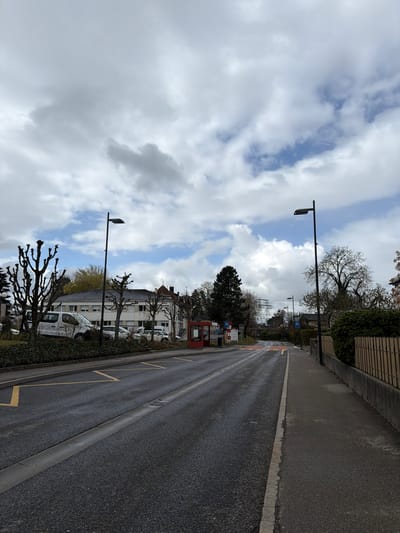 The image depicts an unoccupied street scene in Givisiez, Switzerland, observed from a low, slightly elevated perspective looking down a curved road. The sky above is partially overcast with scattered white and grey clouds, revealing patches of blue sky.
In the foreground, the asphalt road surface appears damp, exhibiting a dark grey color. A narrow concrete median/drainage channel runs longitudinally along the center of the visible road. On the left side of the road, yellow chevron-style markings are present. Further down the road, a yellow and white striped crosswalk is visible. A concrete sidewalk borders the right side of the road, separated by a curb, and continues into the distance. To the right of the sidewalk, a low concrete wall supports a wooden picket fence, behind which dense green hedges and leafless deciduous trees are visible.
On the left side of the street, beyond a small, green-covered embankment, several vehicles are parked, including a white commercial van and multiple cars. A row of bare, pollarded trees with gnarled branches lines this side of the road. Behind these trees and parked vehicles stand multi-story buildings with light-colored facades and dark roofs, some featuring small dormer windows. One building exhibits a distinct reddish roof section. A bright red telephone booth with a clear glass front is positioned on the left, near the road.
Two tall, black, modern streetlights with rectangular LED fixtures are prominent on either side of the road in the mid-ground. In the distance, on the right side of the image, a large metal lattice electrical transmission tower is visible against the sky. Various road signs, including blue rectangular signs with white directional arrows, are sparsely distributed along the road's edge further into the background. No individuals or active interactions are present in the scene.