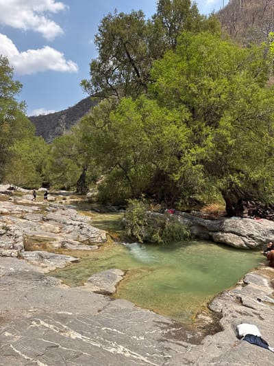 The image depicts a natural riverine environment within San Miguel Atlapulco, Mexico, under a clear blue sky with scattered white cumulus clouds. The foreground consists of large, light-colored stratified rock formations, which form ledges and shallow pools containing clear, greenish-tinted water. In the lower right foreground, a white garment and dark apparel are placed on the rocks.
A stream with light green, flowing water occupies the midground, traversing the rocky terrain and forming small cascades and interconnected pools. On the left bank of the stream, two individuals in dark clothing are walking on the exposed rocks towards the background. On the right bank, a large tree with dense, vibrant green foliage dominates the scene. Beneath this tree, at the water's edge, a person with dark hair, wearing brown attire, is seated on a rock, with their lower legs submerged in the water. Several small, colorful items are visible on the ground near the base of the large tree on the right.
The background features steep, arid-appearing hills or mountains covered with sparse vegetation, including what appear to be cacti and some bare tree trunks. Other green-leaved trees are scattered along the riverbanks extending into the mid-distance. The overall lighting suggests a bright, sunny daytime setting.