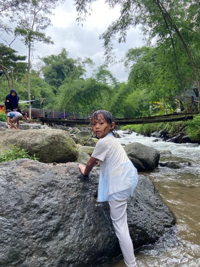 A young girl, wet and clad in white, precariously climbs on a large rock alongside a flowing river. Her gaze is directed towards the camera. Lush greenery, including bamboo, surrounds the area, with a glimpse of a bridge or walkway in the background. Other people are visible near the riverbank. The overall scene suggests an outdoor adventure or recreational activity.