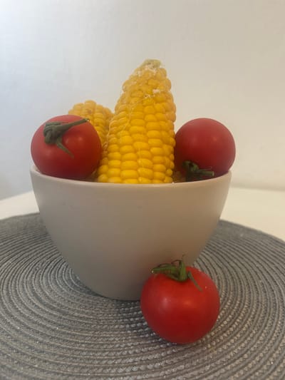 This is a close-up, eye-level shot of a light-colored bowl filled with two cobs of corn and three cherry tomatoes, placed on a circular gray placemat. The corn is yellow and appears to be seasoned with salt. Two of the tomatoes are resting on top of the corn inside the bowl, while the third tomato is positioned in front of the bowl on the placemat. The background is a plain white wall. There are no people or animals in the image. The scene is indoors, likely on a table. The location is not discernible from the visual information provided, and therefore cannot be specified as Banská Bystrica, Slovakia.