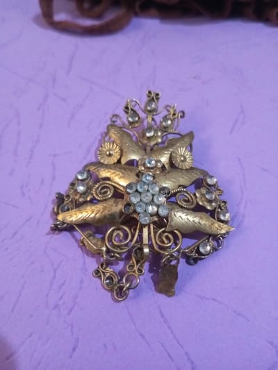 An ornate, golden-colored metal artifact is centrally positioned on a wrinkled, light purple textile surface. This elaborate piece of jewelry features a symmetrical design with a prominent central cluster of multiple small, light-colored, round faceted stones, possibly clear or light blue crystals. Surrounding this central cluster are stylized metallic leaves and smaller floral elements, each typically set with individual similar stones. Intricate scrollwork, filigree, and small, linked chains with flat, rectangular metal charms are integral to the design, extending from the lower portion. The metal exhibits varying shades of gold, with darker patination visible in recesses, indicating age. A blurred, dark brown, textured object is partially visible in the upper background. No individuals are present. This item is located in Chandragiri, Nepal.