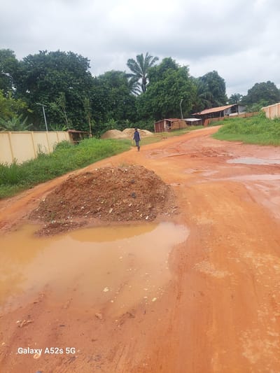 A person walks down a muddy, red dirt road in Umudioka, Nigeria, under a cloudy sky. The road is lined with lush green vegetation and trees. Several small buildings with corrugated metal roofs are visible in the background, along with piles of sand. A large puddle of muddy water reflects the overcast sky in the foreground, next to a mound of reddish-brown dirt and debris. The "Galaxy A52s 5G" watermark is present at the bottom left of the image.