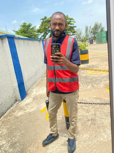 This media file shows a man in Kinkinau, Nigeria, taking a selfie in what appears to be an outdoor industrial or storage area. The man is wearing a navy blue shirt, a red and grey safety vest, tan trousers, and dark blue sneakers. He has a beard and is looking at the camera. In his right hand, he is holding a smartphone to take the picture.
The background shows a concrete ground with yellow markings and black and yellow bollards. There are also chains visible, possibly as barriers. In the distance, there are stacks of green boxes, trees, and a bright sky, suggesting it is daytime with clear weather. The man's posture and the setting imply he might be a worker in this location. The image is oriented vertically, with the top of the man's head to the left and his feet to the right.