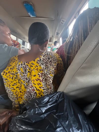 The image shows two women inside what appears to be a bus or van, with luggage and a black trash bag also present. One woman in the foreground is wearing a yellow and black leopard print top and a black braided head covering. She is looking downwards, with her face partially obscured by her hand. The other woman is visible in the lower left corner, looking upwards with her mouth open. She is wearing a gray top. The interior of the vehicle is visible, including seats, a window, and light fixtures on the ceiling. The lighting suggests it might be daytime, possibly overcast, as the interior is somewhat dim. The scene appears to be in motion, given the context of being inside a vehicle. The luggage and trash bag suggest travel or the aftermath of a journey. There are no visible signs of text in the image. The setting is Kugbo, Nigeria.