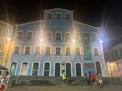 This nighttime image documents the illuminated facade of the Fundação Casa de Jorge Amado, situated in the Pelourinho district of Salvador, Bahia, Brazil. The three-story structure is painted light blue with white trim, featuring numerous white-grid windows across its upper floors and seven dark green arched ground-floor entrances. The building's name is inscribed centrally above the lower doors. A large green banner displaying "QUARTA FREE" is affixed vertically to the upper right facade, and an artistic relief sculpture is present on the lower right wall. Two luminous yellow streetlights are mounted on the building.
A wide, shallow flight of stone steps leads from a rough, unpaved plaza up to the building's entrance level. On the left, two adult females are positioned near a dark cylindrical trash bin, one partially obscured. On the right, a cluster of five to six adults is observed at the base of the steps; one male in a yellow-green shirt and white hat is seated, while the others stand facing each other, conversing. Adjacent structures include a yellow building on the left and a lighter-toned building with arched windows in the background on the right.