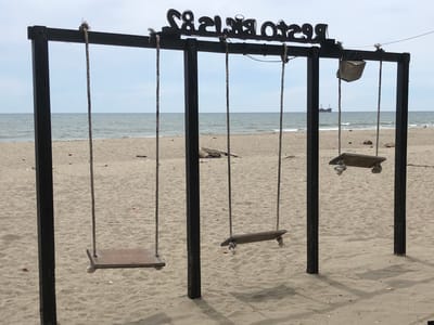 This media file depicts a swingset on a sandy beach with the ocean in the background. The swingset is constructed of black metal posts and a crossbeam. Three wooden swings are suspended by ropes. One swing on the far right appears to be made from a single plank of wood, while the other two are more traditional plank seats. Above the swings, a black sign with white lettering is affixed to the crossbeam. The lettering is in a decorative font and reads "RESTAURANT & BAR." In the distance, a large ship is visible on the water. The sky is overcast, suggesting a cloudy day. The sand is light brown and shows some footprints and debris. The overall scene conveys a relaxed, beachside atmosphere. The location is identified as Keudekruenggeukueh, Indonesia. There are no people visible in the media file. The time of day appears to be daytime due to the natural light illuminating the scene.