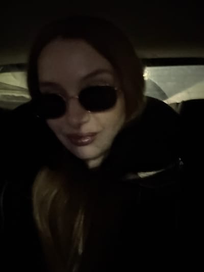 A single individual, an adult female with long, light brown hair, is depicted in a close-up, slightly upward-angled shot. She wears dark, oval-shaped sunglasses with thin metal frames and a dark, possibly black, hooded jacket or coat. Her lips are visible, coated with a glossy, light red or pink lipstick, and she exhibits a slight smile. The scene is dimly lit, suggesting nighttime or a low-light interior environment, possibly inside a vehicle due to the indistinct background features, which include what appears to be a blurred window or light source in the upper right. No visual information within the image definitively identifies the location as Sozina, Montenegro. The individual is looking directly towards the camera, suggesting a selfie or a posed photograph.