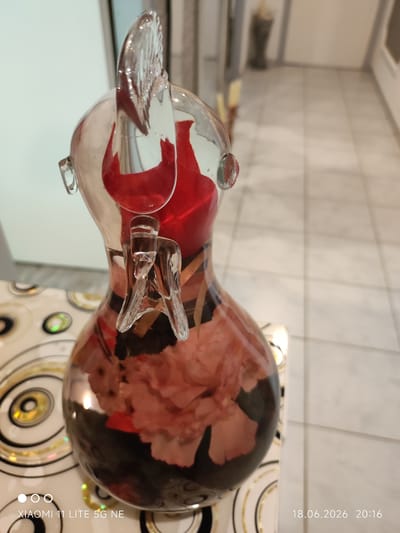 This is a close-up, high-angle shot of a glass figurine, possibly a bird or animal, with a red interior and what appears to be dried flowers or decorative elements inside its translucent body. The figurine is positioned on a patterned tablecloth with concentric circles. In the background, a blurred tiled floor and a wall are visible, along with hints of indoor architecture. The image also contains watermarks for "XIAOMI 11 LITE 5G NE" and the date and time "18.06.2026 20:16." There are no people present in the image. The scene is indoors and the location is not specified to be Böblingen, Germany.