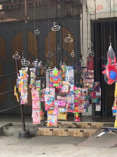 The image shows a street scene in Muliengmanyang, Indonesia, featuring a market stall with an array of colorful toys displayed for sale. The stall is set up outdoors, likely on a sidewalk or in front of a building. Numerous toys are hung from strings and wires, creating a dense display of plastic goods. These toys appear to be aimed at children and include items like toy buckets, toy food, building blocks, and various other small play items. Some toys are packaged in plastic, while others are loose. The background reveals a building with a large, weathered metal door, and beyond that, another entrance with vertical bars. A part of a motorcycle is visible in the lower right corner of the frame, suggesting its presence on the street. The ground is paved, with some stairs leading up to the building. The overall impression is that of a bustling local market or a street vendor preparing for or conducting business.