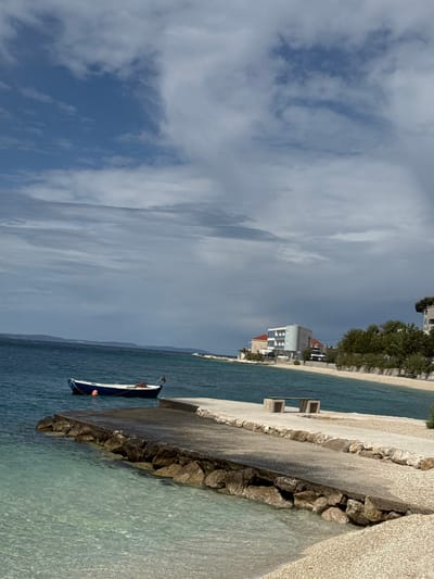 The image shows a view of the Adriatic Sea, taken from a small concrete pier in Dugi Rat, Croatia (latitude: 43.446, longitude: 16.634). The pier is empty, but there is a small blue boat moored to the pier, just off-center, to the left of the image. The sea is a clear, turquoise color. In the background, there is a small beach and a few buildings. The sky is partly cloudy, with streaks of white clouds against a blue sky. The scene is peaceful and tranquil, with no people present. The beach is composed of white, pebbled stones. There is a low-lying concrete wall along the shoreline. Behind the wall, there are some trees lining the shore, and some small buildings are visible on the far shoreline. There are two empty concrete benches placed side-by-side on the beach. A small buoy is visible, just in front of the boat. There is a slight breeze blowing towards the shore. The day is sunny, with a light breeze, and clear skies.