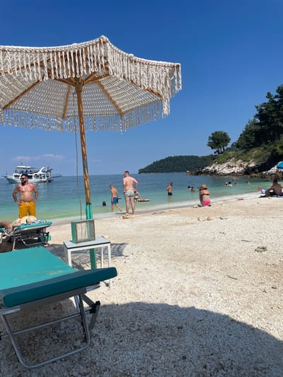 This image depicts a sunny day at a pebble beach in Thassos, Greece. In the foreground, a white macrame umbrella with fringe shades a turquoise lounge chair. To the right of the umbrella is a small white table with a turquoise box on top. The beach is covered in white pebbles, and the turquoise water of the Aegean Sea is visible in the background.
Several people are enjoying the beach and water. A man in yellow shorts stands near the lounge chair, facing the sea. Further back, a group of people are in the shallow water, including a boy in blue swim trunks and a man in grey swim trunks. Another man is sitting on the beach to the right, partially shaded by an umbrella. A white boat with several people on deck is anchored in the bay. The coastline is lined with lush green trees and rocky outcrops. The sky is a clear, bright blue.