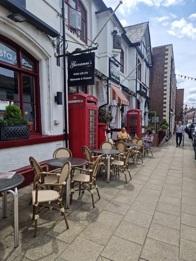 The image captures a street scene in Knutsford, United Kingdom, likely on a sunny day given the shadows. In the foreground, an outdoor seating area for a restaurant named "Giovanni's Ristorante & Pizzeria" is set up on a paved sidewalk. Several tables and chairs made of rattan and metal are visible.
A distinctive red British telephone box stands next to the restaurant's entrance, with another one further down the street. The restaurant itself is a white building with dark red window frames and a dark awning. A sign for "Giovanni's" with a phone number and description hangs above the entrance.
Two patrons are seated at a table outside the restaurant: a man in a yellow t-shirt and a woman in a floral dress. Further down the street, a man in a light blue shirt and dark trousers walks away from the camera. The street is lined with other buildings, including a brick structure with decorative stone detailing. Cars are parked along the right side of the street, and a line of colorful bunting stretches across the street in the distance. The sky is partly cloudy.