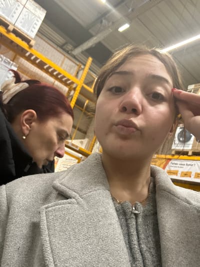 A selfie-style photo captures two women in what appears to be an indoor commercial or industrial setting, likely a warehouse or large retail store. The image is taken at an upward, tilted angle.

In the foreground, a woman's face is prominent, looking directly into the camera. She has light brown hair, and her left hand is partially visible, with a finger touching her forehead. Her lips are slightly pursed, and her expression is somewhat neutral. She is wearing a light grey, textured coat with large lapels and a visible pocket flap on the right side. A thin gold necklace is also visible around her neck.

In the mid-ground and background, the profile of a second woman is visible. She has reddish-brown hair tied back, possibly with a light-colored hair claw or clip visible at the back of her head. She is wearing a dark, possibly black, jacket or coat, and a small gold stud earring can be seen in her right ear. She is looking towards the left side of the frame, away from the camera.

The background consists of typical warehouse or store infrastructure. Bright yellow metal shelving units are clearly visible, stretching into the distance. On these shelves, white, shrink-wrapped packages or pallets are stacked, particularly in the lower left portion of the image. More boxed products are visible on shelves in the upper right. Overhead, long fluorescent strip lights illuminate the space.

Part of a French phrase, "Faites-vous livrer!" (meaning "Get it delivered!"), is visible on a box on one of the shelves in the upper right quadrant of the image. The overall impression is that of a casual moment captured in a functional, commercial environment.