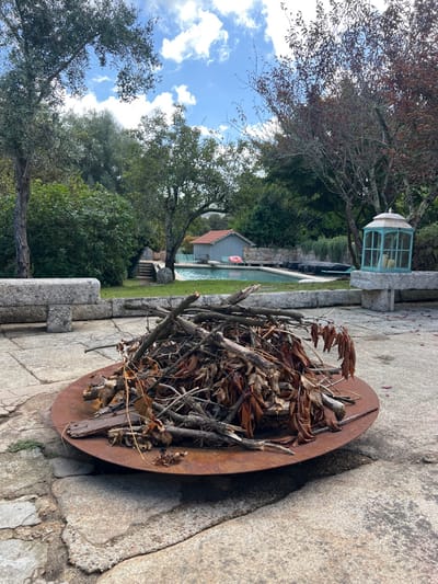 The image depicts an outdoor recreational area in Vila Nova de Cerveira, Portugal, under a partly cloudy blue sky. In the immediate foreground, a round, rusty metal fire pit is situated on a patio composed of large, irregularly shaped grey stone pavers. The fire pit is filled with a dense pile of dry kindling, consisting of numerous branches, sticks, and dried brown leaves.
Beyond the fire pit, a landscaped area unfolds. To the left, a long, rough-hewn grey stone bench is visible. To the right, another similar stone bench supports a large, teal-framed lantern with glass panels, containing three white, cylindrical objects resembling candles.
In the midground, a light blue swimming pool is visible, bordered by a paved deck. Two lounge chairs with striped covers are positioned next to the pool. A pink inflatable ring is either floating in the pool or resting at its edge. Behind the pool, a small, light grey wooden building with a distinctive red-tiled roof is situated.
The background and sides of the scene are densely vegetated with various trees and shrubs. Some trees display green foliage, while others exhibit reddish-brown leaves. The overall environment suggests a private or resort setting. No individuals are present in the frame.
