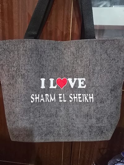 The image shows a close-up of a gray tote bag with white text and a red heart embroidered on it. The text reads "I LOVE SHARM EL SHEIKH". The bag appears to be made of a textured fabric. A black strap is visible on the left side of the bag. The bag is lying on a dark, wooden surface, possibly a table. The lighting suggests it is daytime, and the setting is likely indoors. There are no people visible in the image. The context provided for Karasu, Uzbekistan, is not directly evident from the visual content of the image itself. The overall impression is of a souvenir item or promotional merchandise.
