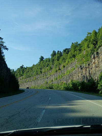 The image depicts a scenic drive through a rocky, forested landscape. The perspective is from inside a vehicle, looking out towards a steep, layered rock cliff face covered in lush green vegetation. A paved road with yellow and white lane markings curves into the distance, shadowed by the cliffs and trees. The sky is a clear, bright blue with a few wispy white clouds, suggesting a sunny day, likely in the late morning or early afternoon. The overall impression is one of natural beauty and a peaceful journey. There is a dark-colored motorcycle visible on the road in the distance. The location is identified as Berea, United States.