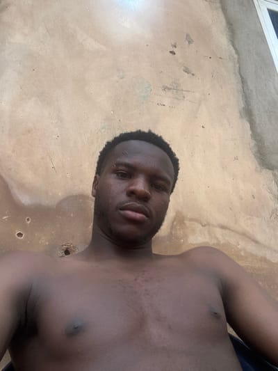 A shirtless, dark-skinned man lies on his back, facing the camera. He has short, dark hair and a slight beard. His eyes are open, and his lips are slightly parted. He is lying on a textured, light brown surface, possibly concrete, with some stains or irregularities. Part of a window is seen in the upper left corner.