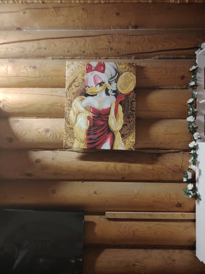 A framed painting of Daisy Duck dressed in a red gown and a gold-colored fur stole, holding a hand mirror, is hung on a log wall. The painting is centrally located and appears to be the main subject. To the right of the painting, a string of artificial white flowers with green leaves is draped vertically down the wall. In the lower left portion of the image, a black object, possibly a television or a cabinet, is visible. The background is a wall constructed from horizontal wooden logs. The image was taken in Gorshki, Russia.