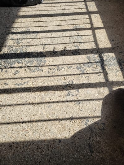 The scene depicts an outdoor ground surface in Ανδραβίδα, Greece, illuminated by direct sunlight. The ground is a light-colored, worn concrete or asphalt pavement, exhibiting darker, irregular stains and visible aggregate. Sharp, elongated, parallel shadows, consistent with a grid or bar-like structure, are cast across the surface. A large, amorphous dark shadow, likely from a nearby object or individual, occupies the bottom right quarter of the frame. A dark, indistinct object, possibly a bag or tire, is partially visible at the top-left edge. No individuals are clearly identifiable, and no specific actions or interactions are underway.