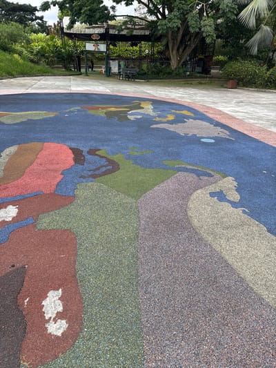 The image displays an outdoor recreational area, specifically the "Mapa Mundial" section within Bioparque Ukumarí, Pereira, Risaralda, AMCO, Colombia. The foreground is dominated by a large, circular, multi-colored map of the world rendered on a rubberized ground surface. Continents are depicted in shades of red, green, brown, light blue, grey, and yellow, contrasting with dark blue sections representing oceans. Visible wear is present on several areas of the map.
A light-colored concrete pathway separates the map from the midground. In the midground, a partially visible covered structure with a dark, possibly thatched roof functions as an entrance or kiosk. A sign featuring a stylized, multicolored bird graphic is mounted on this structure. One individual, attired in dark clothing, is observed standing near the entrance. A dark-colored bench is positioned on the concrete path to the right of the entrance. The background consists of dense tropical vegetation and large trees under an overcast sky.