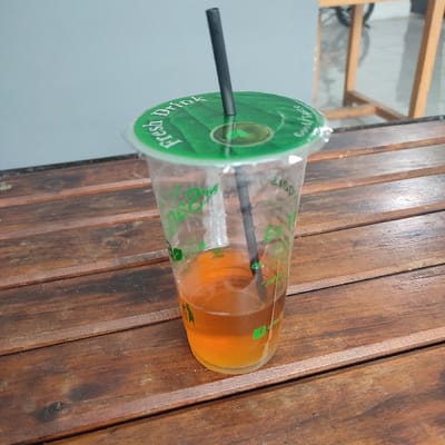 A clear plastic beverage cup, approximately one-third full with an amber-colored liquid, rests on a dark reddish-brown slatted wooden table. The cup features green leaf-shaped logos and the text "Fresh Drink" and "Good Taste" in green lettering. It is sealed with a green plastic film lid, also printed with "Fresh Drink" and a leaf motif, through which a single black plastic straw is inserted. The background reveals a plain light grey wall, and to the right, a partial view of a wooden furniture structure and a metallic object, possibly a bicycle component, are visible. No individuals are present. While the specific address is not discernible from the image, the scene is consistent with a casual eatery or cafe environment in Sukabumi, Indonesia.