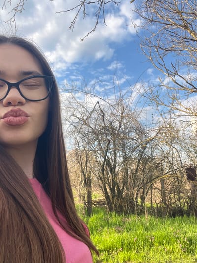 The image depicts a low-angle view of a person with long brown hair wearing black-framed glasses and a pink garment. Their eyes are closed. Above them, a blue sky with white clouds is visible through bare tree branches. To the right, there is green grass dotted with small purple flowers. There is no evidence in the image to suggest a location in Blaskovo, Bulgaria.