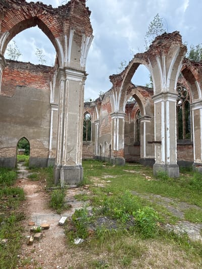 The image depicts the ruins of what appears to be a historical building, possibly a church or a synagogue, located in Bachury, Poland. The structure is characterized by crumbling brick walls, arched openings, and weathered stone columns. Vegetation, including grass and small trees, is growing both inside and outside the ruins, indicating a state of disrepair and abandonment. The sky is overcast. The foreground shows a path made of gravel and debris, with scattered bricks and stones. The scene is static and quiet, conveying a sense of decay and the passage of time. There are no people or animals visible in the image.