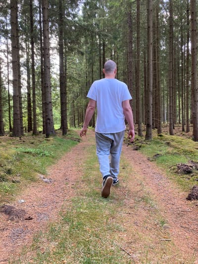 The image, originally oriented sideways, depicts a person from behind walking along a forest path. When viewed correctly (rotated 90 degrees counter-clockwise), the person, with a shaved head, is wearing a plain white t-shirt, grey sweatpants, and black sneakers with white soles. They are positioned towards the center-right of the frame, moving away from the viewer.
The setting is a dense coniferous forest, characterized by numerous tall, slender trees with dark grey trunks and green needles, suggesting spruce or pine trees. Bright sunlight filters through the tree canopy, creating bright spots and a dappled lighting effect, indicating it is daytime. The path itself is light brown, composed of dirt and small pebbles, with areas of green grass and moss growing along its edges. The forest floor on both sides of the path features dark green undergrowth and moss. The scene evokes a peaceful, natural outdoor environment. There is no visible text. The location context is Vessigebro, Sweden.