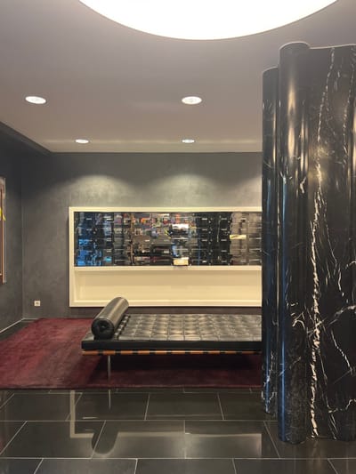 This image depicts a modern interior space, located in Uccle - Ukkel, Belgium. The floor is paved with large, dark grey rectangular tiles exhibiting light reflections. A dark red rectangular rug is positioned on the tiled floor, beneath a black tufted leather Barcelona-style daybed featuring a cylindrical bolster cushion, a wooden slat base, and chrome legs. To the right, a fluted pillar constructed of black marble with prominent white veining extends from floor to ceiling. The far wall, painted in a textured dark grey, hosts a large, white-framed display unit. This unit includes a lower white shelf and an upper section filled with numerous small, reflective, rectangular elements arranged in rows. A partially visible framed artwork with a brown border and yellow accents is on the left wall, above a white electrical outlet. The white ceiling is illuminated by multiple circular recessed spotlights and a large, central curved flush-mounted light fixture. No individuals, actions, or interactions are present.