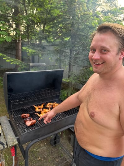 A man is grilling meat outdoors. He is shirtless, wearing black shorts with a blue waistband. He has fair skin, light brown hair, and a light beard. He is smiling at the camera. In front of him is a black charcoal grill with chicken wings and sausages on the grates. The man is pointing at the food with his right hand. The grill is in a backyard with trees and a fence in the background. The ground is covered in dirt and grass. The image appears to be taken in Усть-Мана, Russia, given the presence of coniferous trees common to the Siberian region.