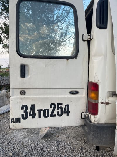 The image shows the rear and side of a white Ford Transit van parked on a gravel lot in Vardanashen, Armenia. The rear doors are open, revealing a reflection of a tree and the sky in the passenger-side window. The van is weathered and shows signs of damage, including rust and dents on the bodywork. The text "AM 34 to 254" is visible in black lettering on the lower portion of the passenger-side rear door. A red and orange taillight is visible on the driver's side rear of the van. The surrounding environment appears rural, with a fence and some vegetation visible in the background.