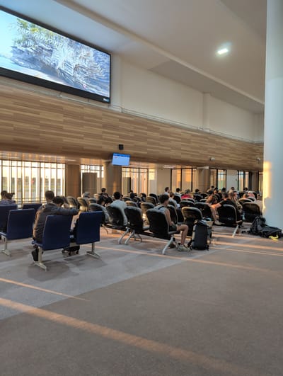 The image depicts a waiting area, likely an airport or transportation hub, with a large screen displaying a landscape image of rocky cliffs and trees. The room is furnished with rows of blue and black seating. Several individuals are seated, facing various directions, some engaged in conversation or looking at devices. Bags and personal belongings are placed near the seats. Sunlight streams in from large windows on the left, casting long shadows across the carpeted floor. The architecture features a wooden-paneled wall and a white ceiling with lighting fixtures. The environment suggests a public waiting space. The sign in the bottom right corner of the large screen indicates "Plan B", a digital signage company. The overall scene is one of people waiting in a modern, well-lit public space.
